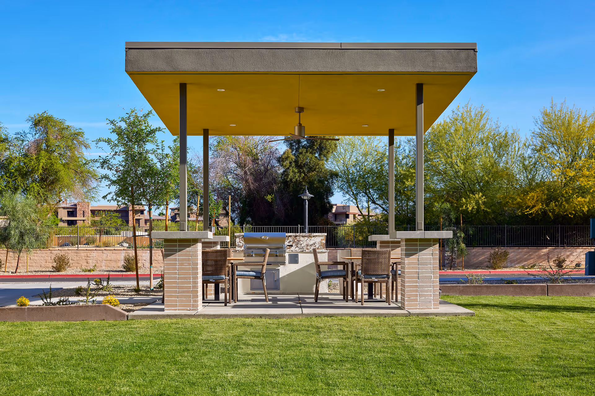 Outdoor covered seating area with a built-in grill and several wicker chairs around tables, set on a concrete patio surrounded by green grass and trees under a clear blue sky.