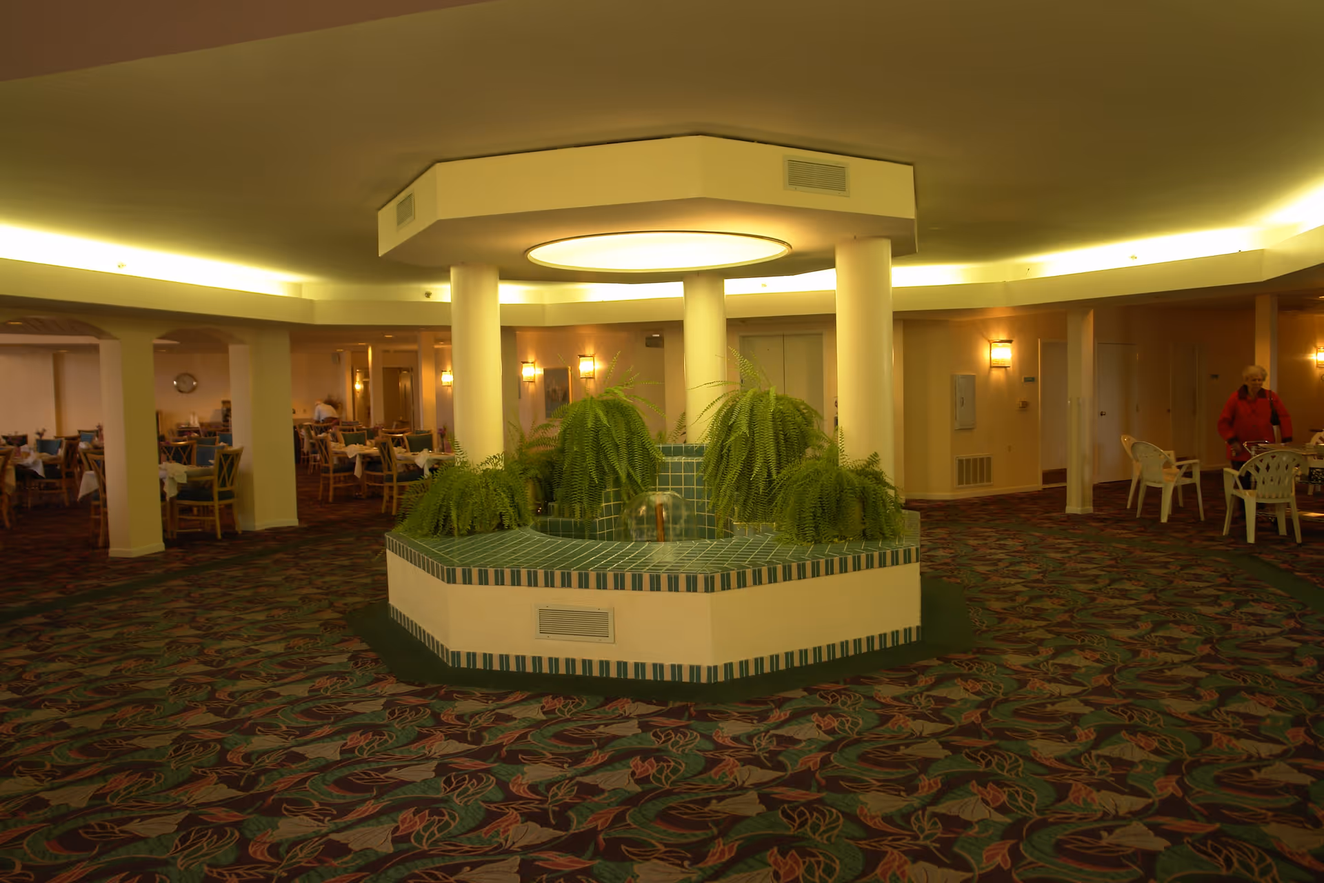 Interior view of a senior living facility with a central planter featuring green ferns surrounded by four white columns and a circular light fixture above. The room has patterned carpet and is furnished with multiple dining tables and chairs. A few people are visible in the background.