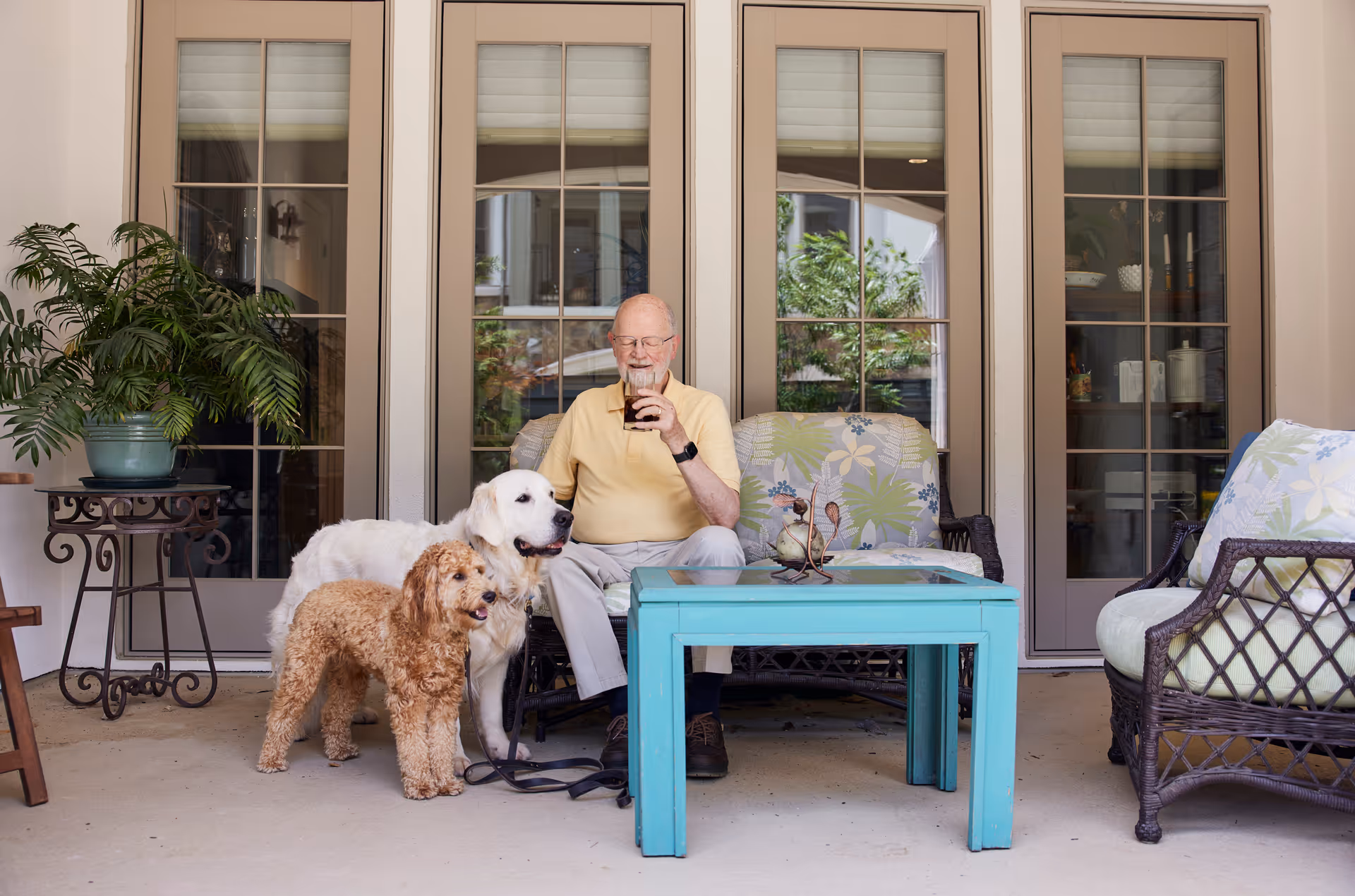 An elderly man sits on a covered patio armchair with two dogs beside him and a turquoise coffee table in front of French doors.