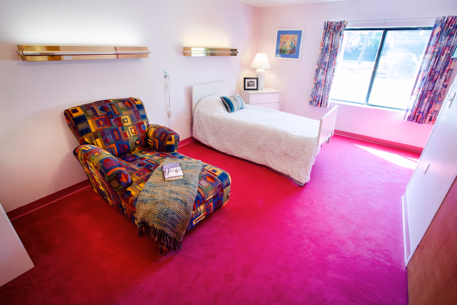 A bright senior living bedroom with a single bed, colorful patterned armchair, bedside table with a lamp, and a large window.