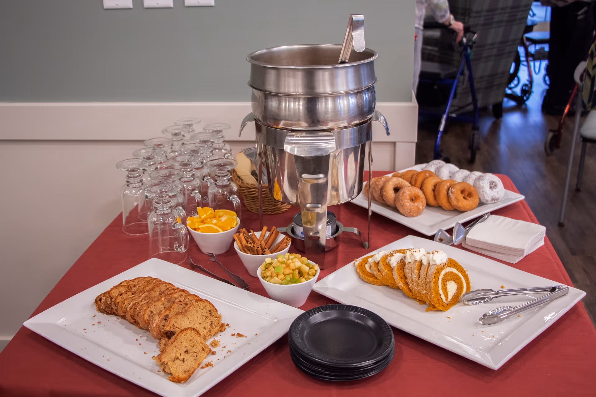 Buffet table in a dining area with a coffee urn, stacked glasses, plates, sliced cakes, donuts and bowls of fruit on a red tablecloth.
