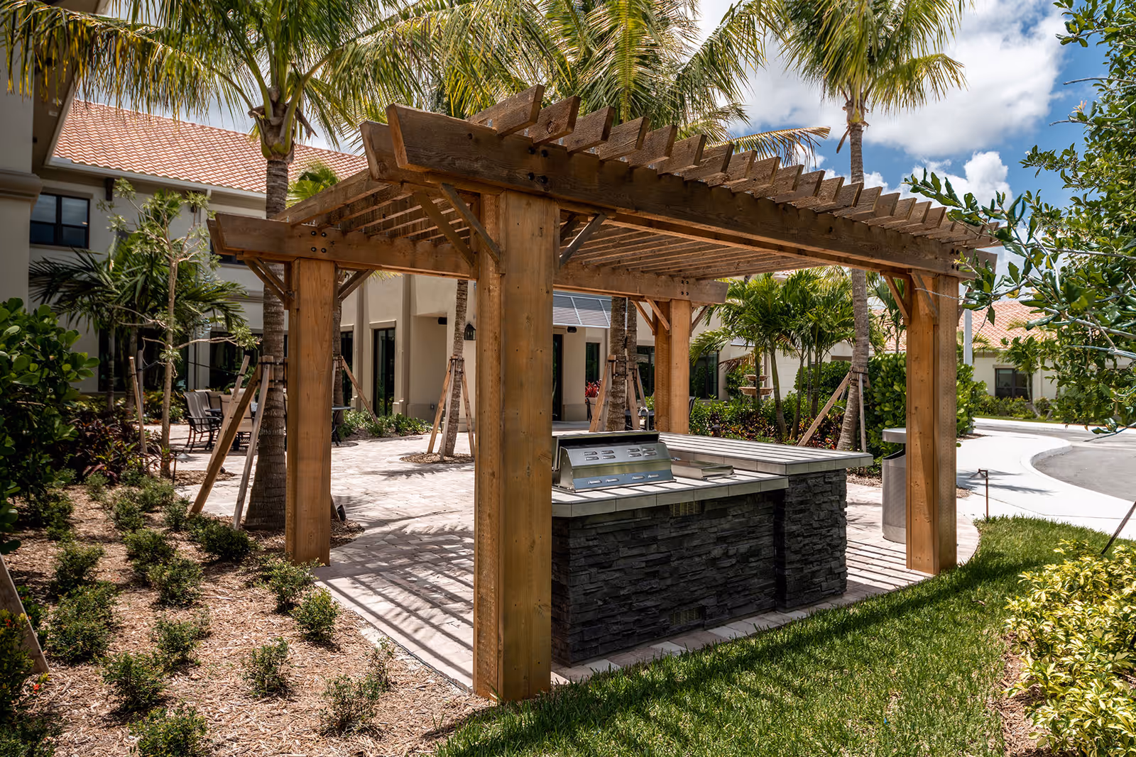 Wooden pergola with a built-in stone-faced outdoor grill and counter in a landscaped courtyard with palm trees and nearby senior living buildings.