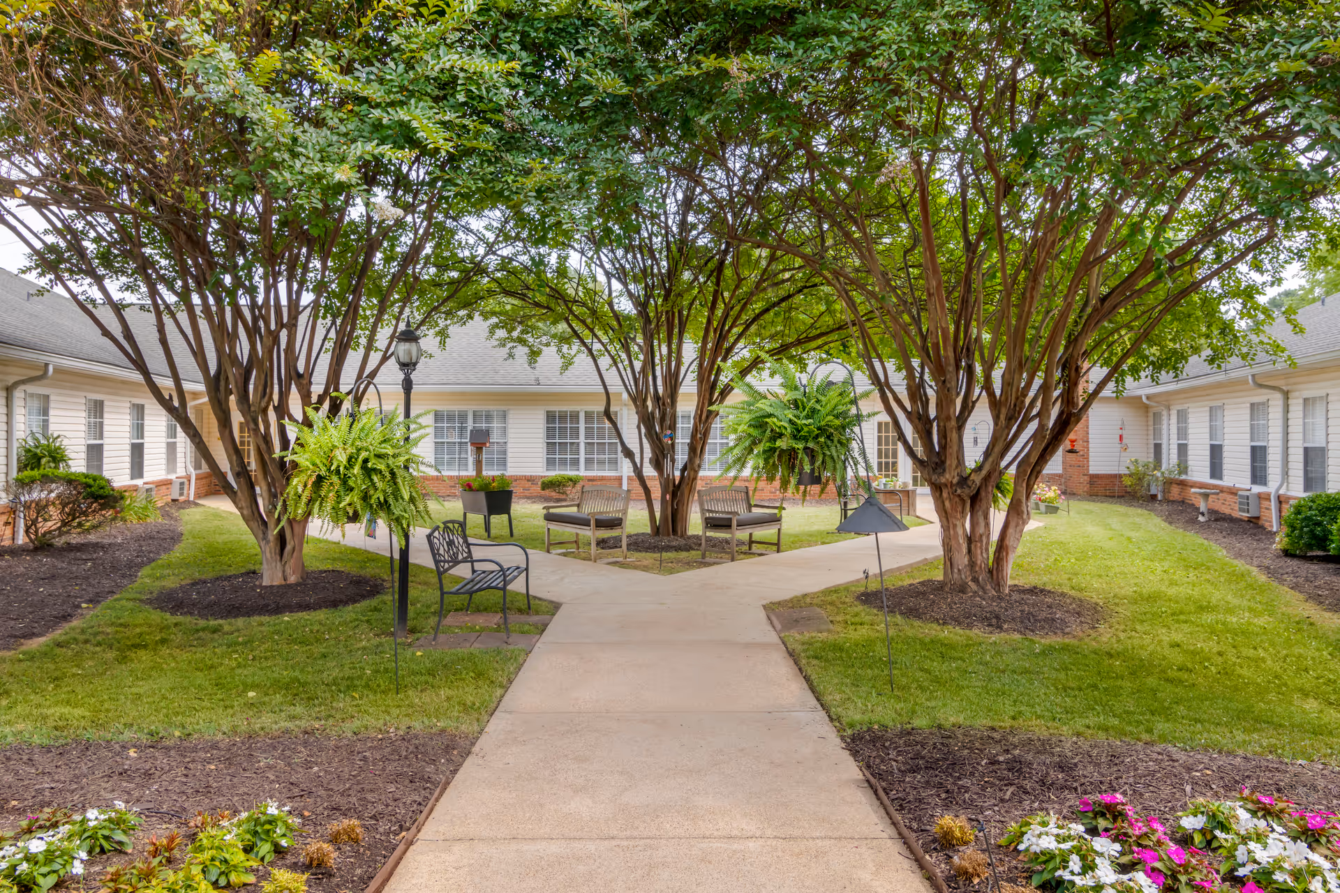 A peaceful outdoor courtyard at Brookdale Hixson featuring a paved walkway that splits into two paths surrounded by green grass, trees, hanging ferns, benches, and flower beds. The courtyard is enclosed by a single-story building with white siding and multiple windows.