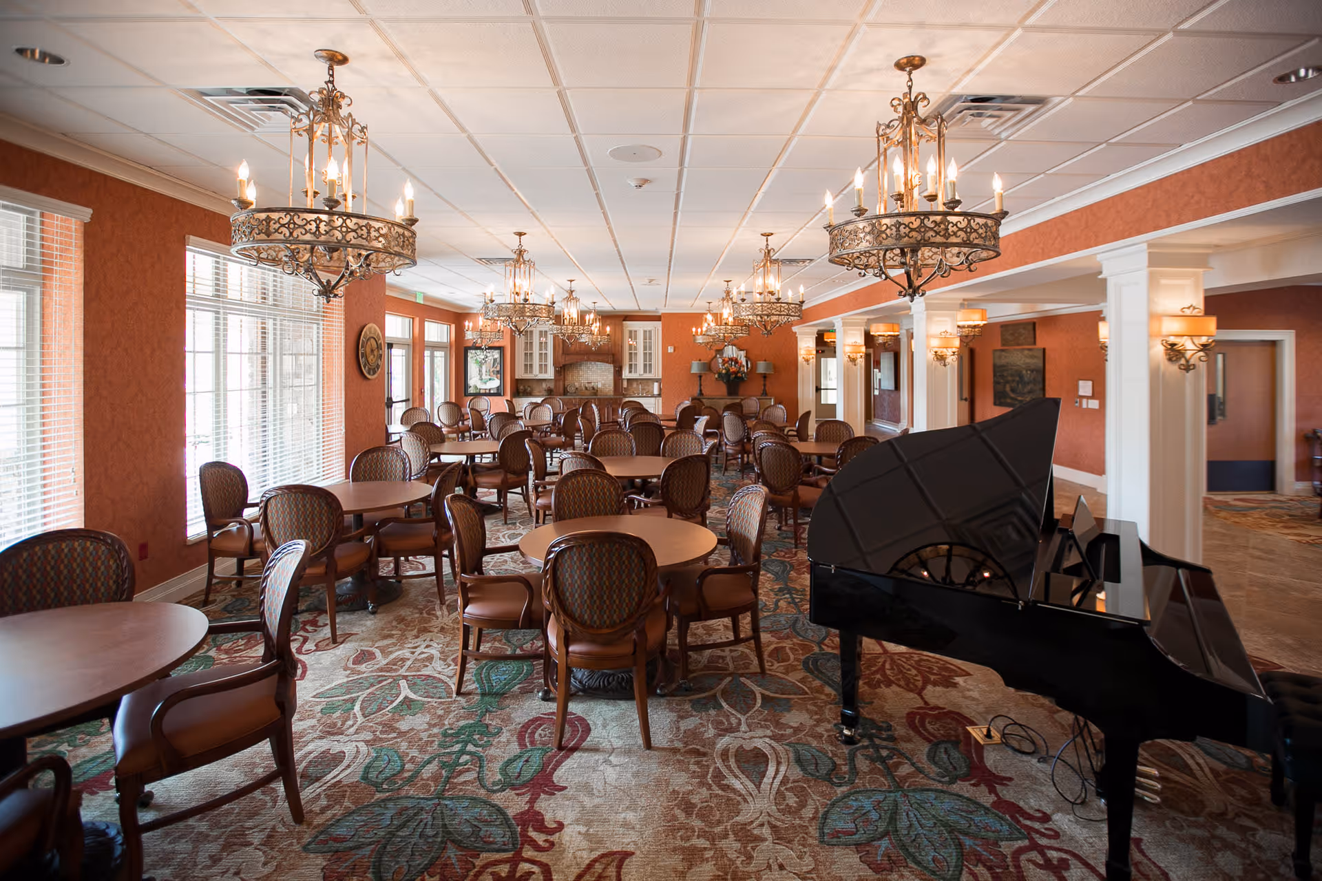 A spacious dining room with multiple round wooden tables and cushioned chairs arranged neatly. The room features ornate chandeliers hanging from the ceiling, large windows with blinds on the left side, and a grand piano on the right. The walls are painted a warm reddish color, and the carpet has a floral pattern. There is a small kitchen or serving area at the far end of the room.