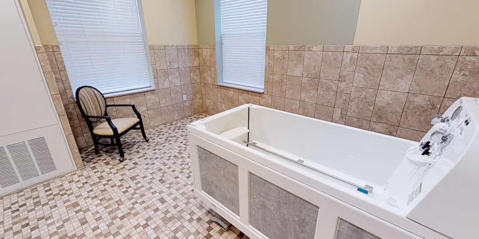 A tiled room with a large white walk-in bathtub on the right side and a cushioned wooden chair with armrests near two windows with closed blinds. The walls are partially tiled with beige tiles and the floor has a mosaic tile pattern.
