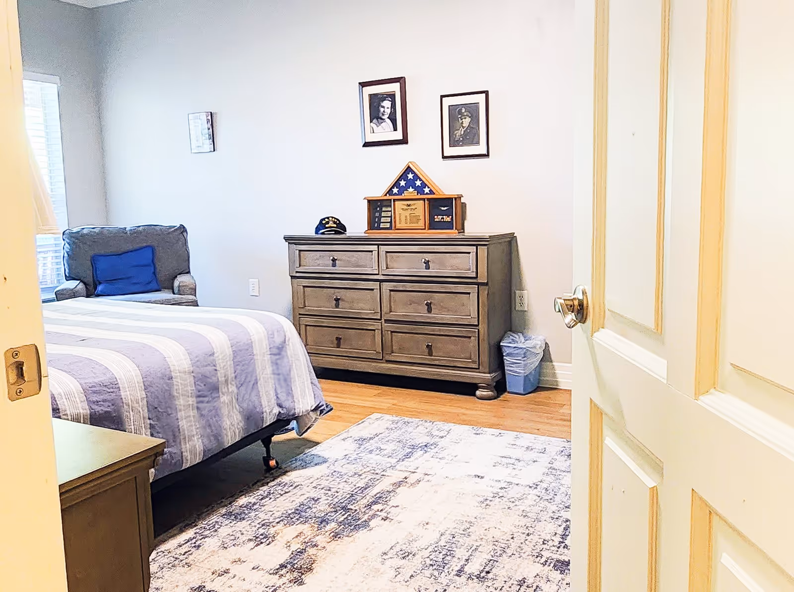 View into a bedroom with a bed covered in a striped purple and white blanket, a gray armchair with a blue pillow near a window, a wooden dresser with six drawers, and framed photos and a folded American flag displayed on the wall above the dresser. A small trash bin is placed next to the dresser on a wooden floor with a patterned area rug.