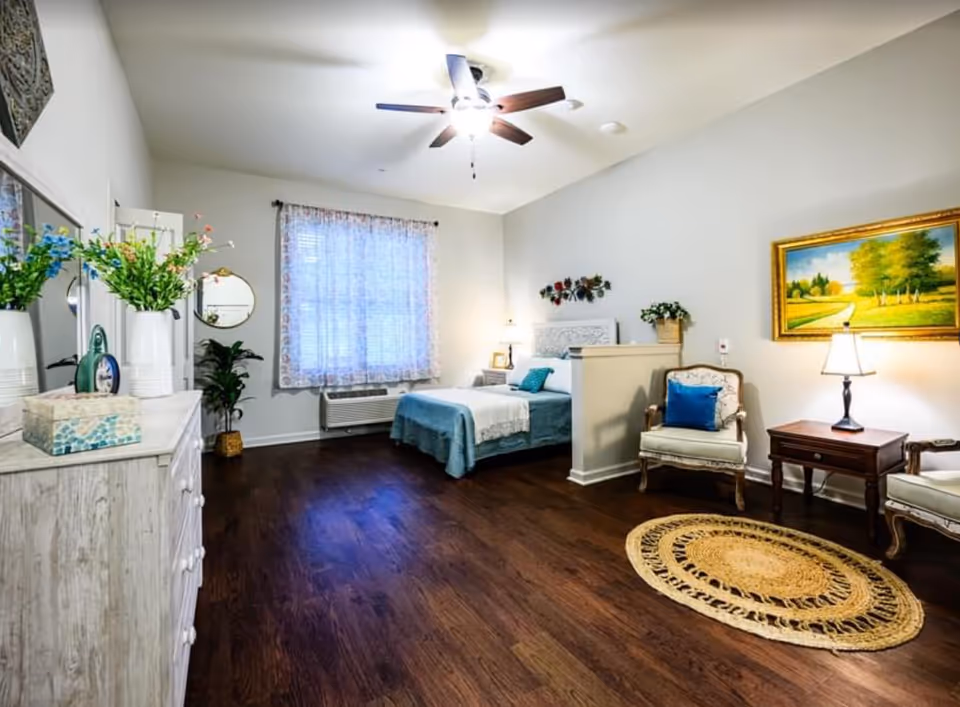 Well-lit furnished senior living bedroom with a bed, seating area, dresser, ceiling fan, and hardwood floors.