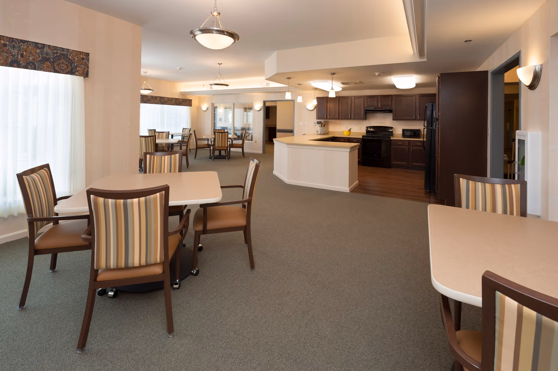 A spacious dining area with multiple tables and chairs featuring striped upholstery. The room has large windows with sheer curtains and valances, and is well-lit with ceiling lights. In the background, there is an open kitchen with dark wood cabinets, a black stove, refrigerator, and a countertop bar area.