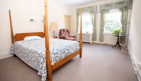 A bedroom with a wooden four-poster bed covered with a blue and white patterned bedspread. There is a brown upholstered armchair next to a floor lamp near a closed door. Two windows with green curtains let in natural light, and a small round table with a plant is placed between the windows. The room has beige carpet and white walls.