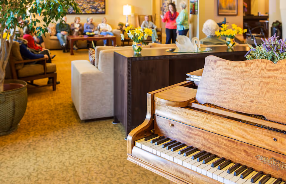 A cozy living room area in a senior living facility with a wooden piano in the foreground. Several elderly people are seated on sofas and chairs in the background, engaging in conversation. The room is warmly lit with lamps and decorated with flowers and paintings on the walls.