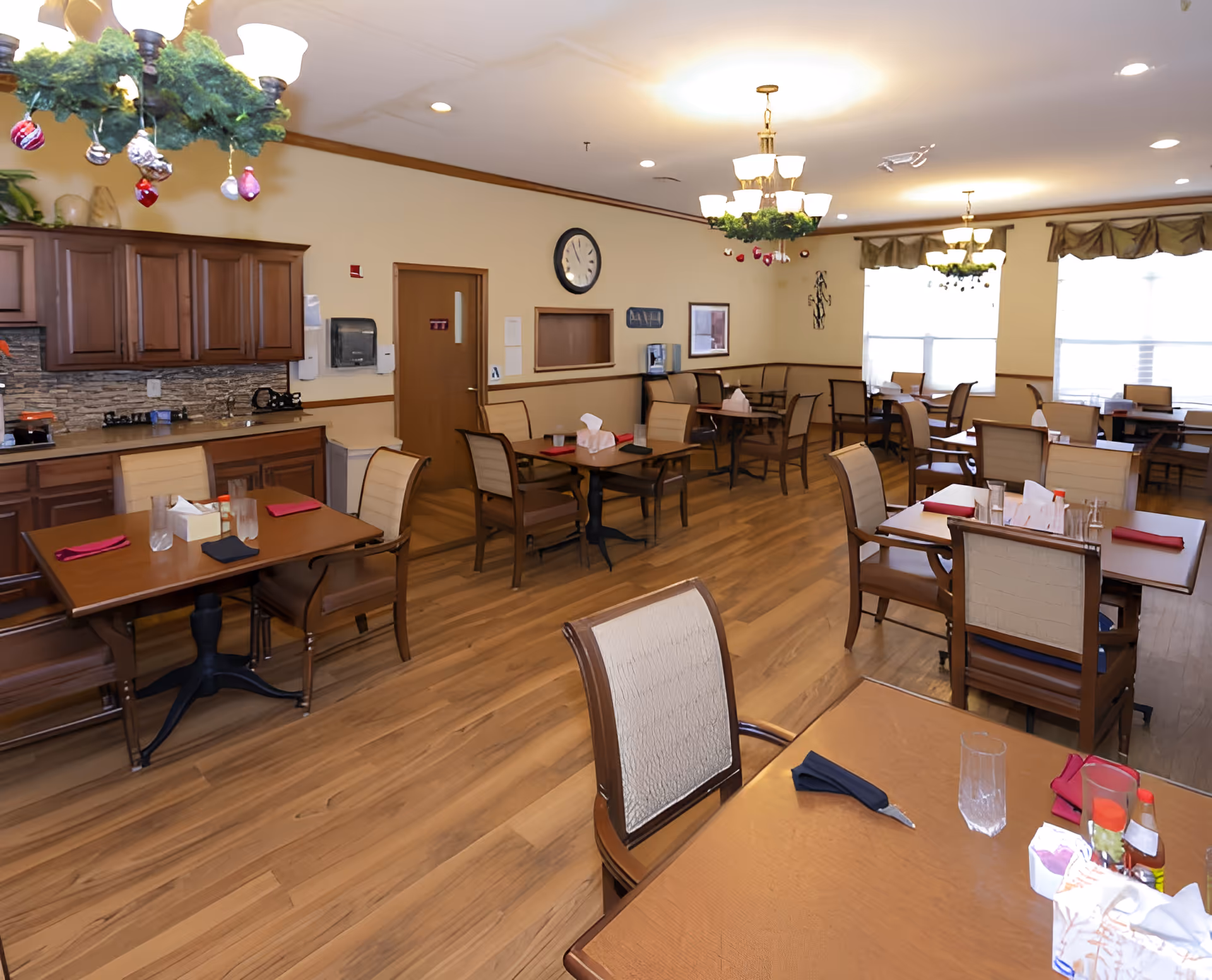 A senior living facility dining room with multiple wooden tables and chairs arranged neatly. The room has wooden flooring, beige walls, and large windows with valances allowing natural light to fill the space. There are chandeliers decorated with greenery and ornaments hanging from the ceiling. A clock is mounted on the wall above a door, and there is a countertop with cabinets and a paper towel dispenser on the left side.