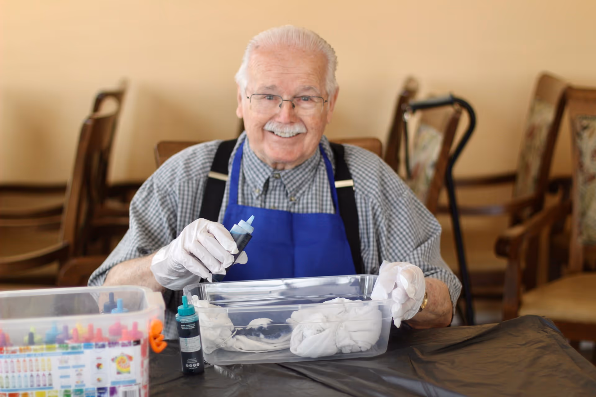 An elderly man wearing glasses, a blue apron, and white gloves is sitting at a table engaged in a tie-dye activity. He is holding a bottle of dye over a plastic container with white fabric inside. There are chairs and a walking cane in the background.