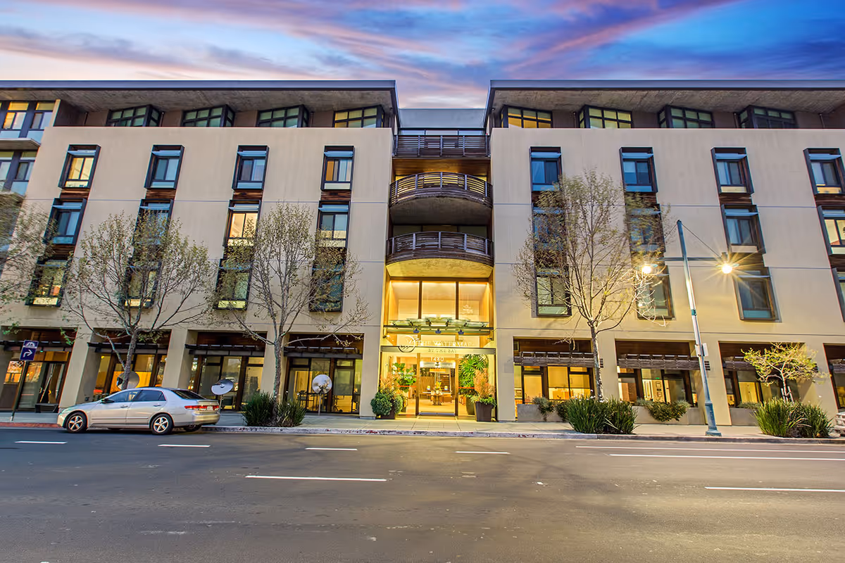 Front exterior of a multi-story senior living building with an illuminated main entrance facing the street at dusk.