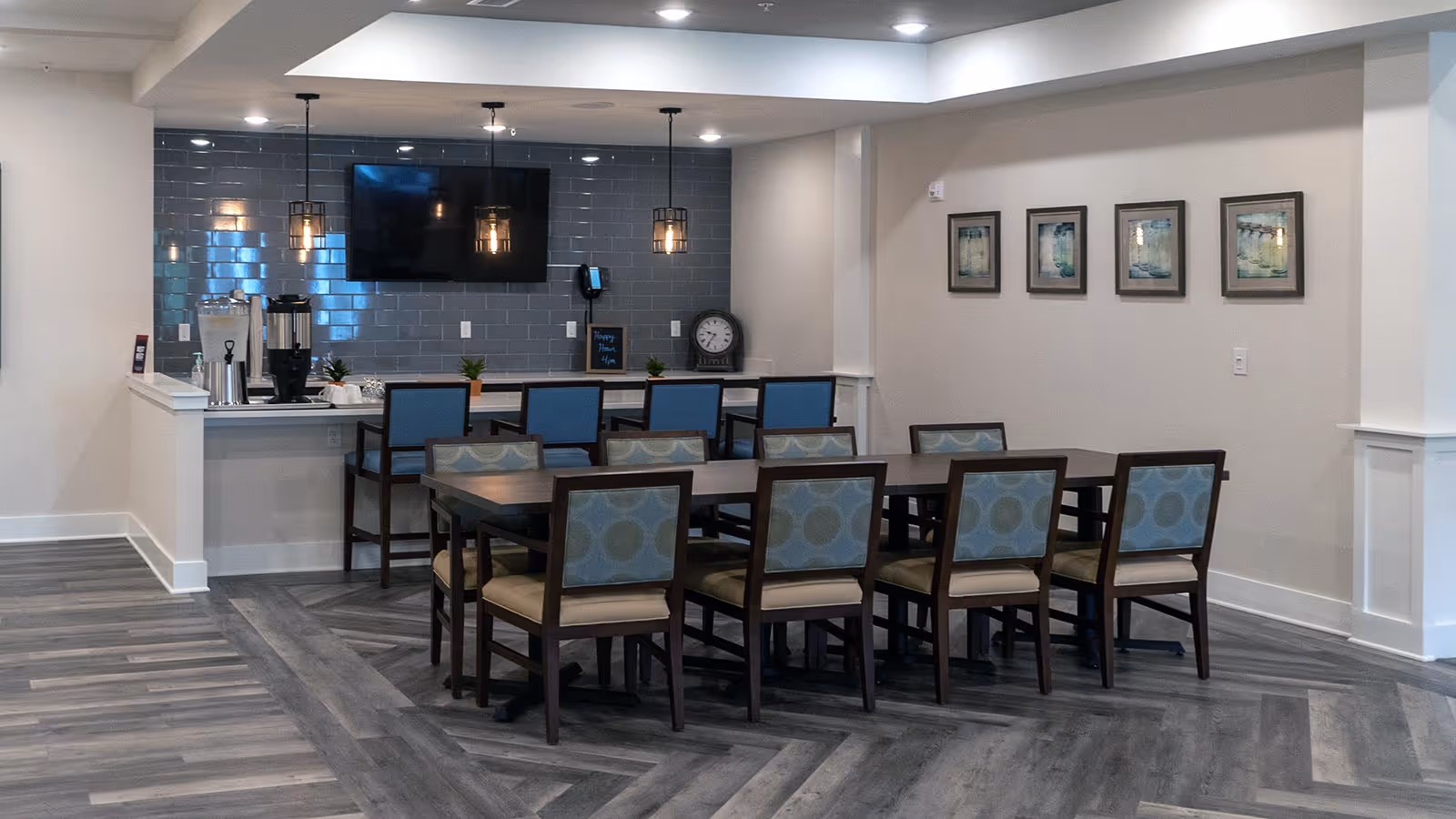 Communal dining area with a rectangular table and chairs, a service counter with a coffee station, pendant lights and a wall-mounted TV.