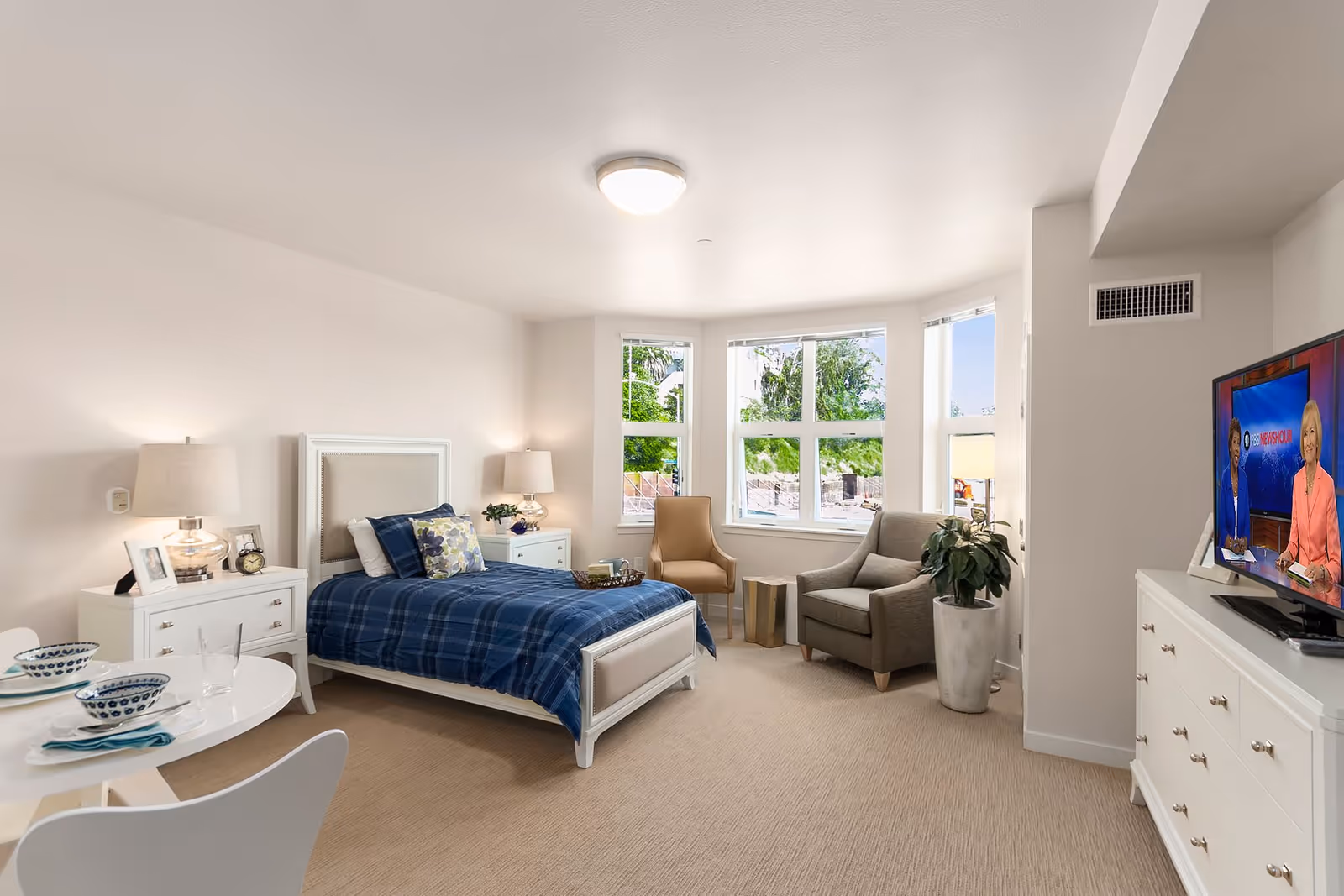 A bright and cozy bedroom in a senior living facility featuring a single bed with blue plaid bedding, two white nightstands with lamps, a small round dining table set for two, a beige armchair and a gray armchair near a large bay window, a potted plant, and a white dresser with a flat-screen TV displaying a news program.