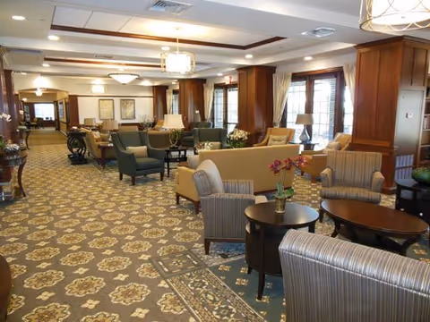 Spacious and well-lit common area in an assisted living facility with patterned carpet, multiple seating arrangements including armchairs and sofas, wooden columns, large windows with curtains, and decorative lighting fixtures on the ceiling.