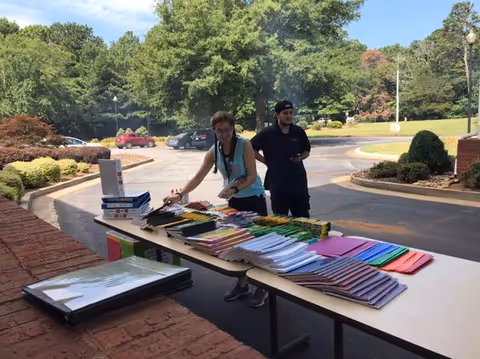 Two people standing outdoors behind a long table covered with neatly arranged school supplies including notebooks, folders, pencils, and crayons. The setting is a parking lot area with trees and bushes in the background under a clear sky.