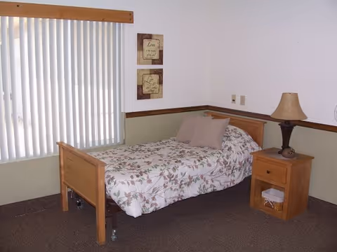 A simple bedroom with a single wooden bed covered with a floral patterned bedspread and two pillows. Next to the bed is a wooden nightstand with a lamp and a basket on the lower shelf. The room has beige walls with a wooden trim and a window with vertical blinds. Two framed decorative wall hangings are above the bed.