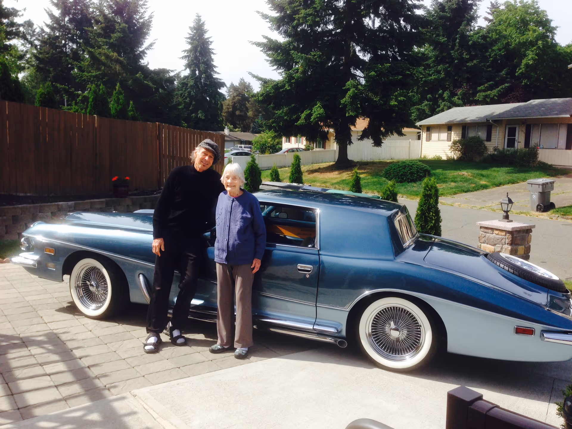 Two older adults stand beside a vintage blue car parked in a residential driveway with houses and trees in the background.
