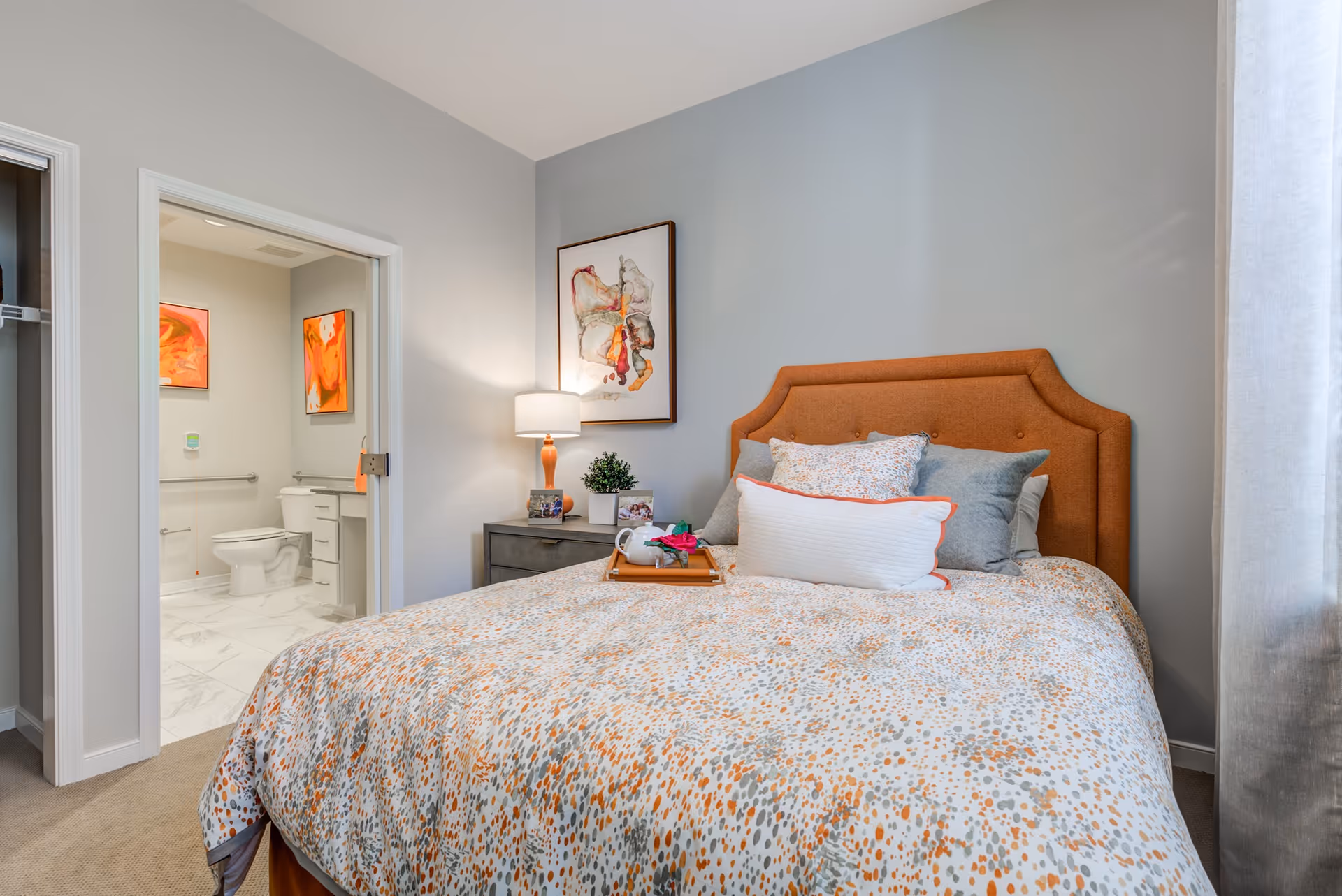 A cozy bedroom with a bed featuring an orange upholstered headboard and a patterned bedspread in shades of orange, gray, and white. There are multiple pillows on the bed, and a wooden tray with a teapot and a flower. Next to the bed is a nightstand with a lamp, a small plant, and framed photos. The wall behind the nightstand has an abstract painting. An open doorway reveals a bathroom with a toilet, vanity, and two orange-themed paintings on the walls.