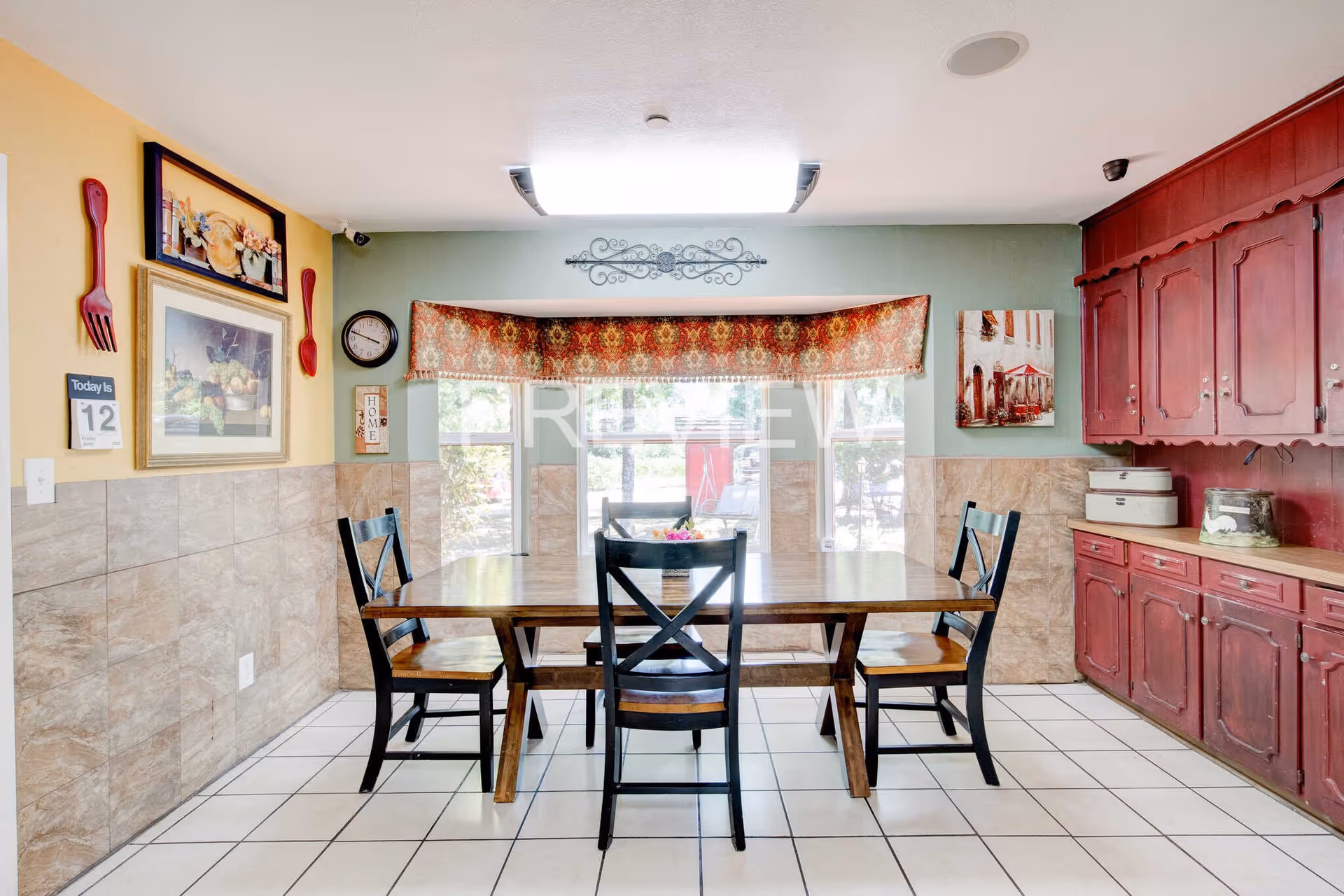 A dining room with a wooden table and four black chairs. The walls are painted yellow and green with tile halfway up. There are decorative items on the walls including framed pictures, a clock, and large red fork and spoon wall hangings. A window with a patterned valance lets in natural light. On the right side, there are red wooden cabinets with countertop storage boxes.