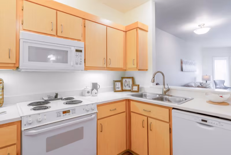 A bright kitchen with light wood cabinets, a white electric stove and oven, a white microwave above the stove, a double stainless steel sink with a modern faucet, and a white dishwasher. The kitchen counter has a few decorative items including framed pictures and a bowl. In the background, a living room area with a couch, lamp, and window is partially visible.