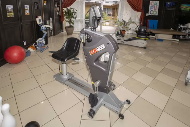 Indoor exercise room with various fitness equipment including a SciFit recumbent exercise bike in the center, a red exercise ball, and other workout machines. The room has tiled flooring, large windows with curtains, and some potted plants near the windows.