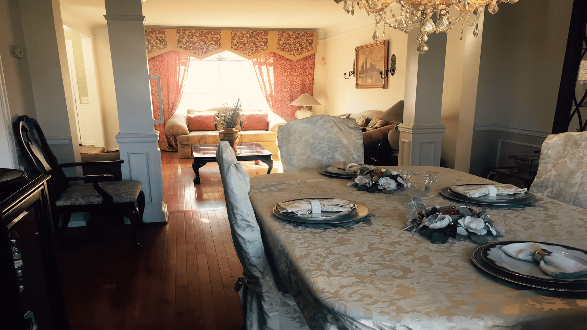 A warmly lit interior space featuring a dining table covered with a patterned tablecloth and set with plates, napkins, and floral decorations. In the background, there is a living room area with a sofa, coffee table, and large window with red patterned curtains allowing natural light to fill the room.