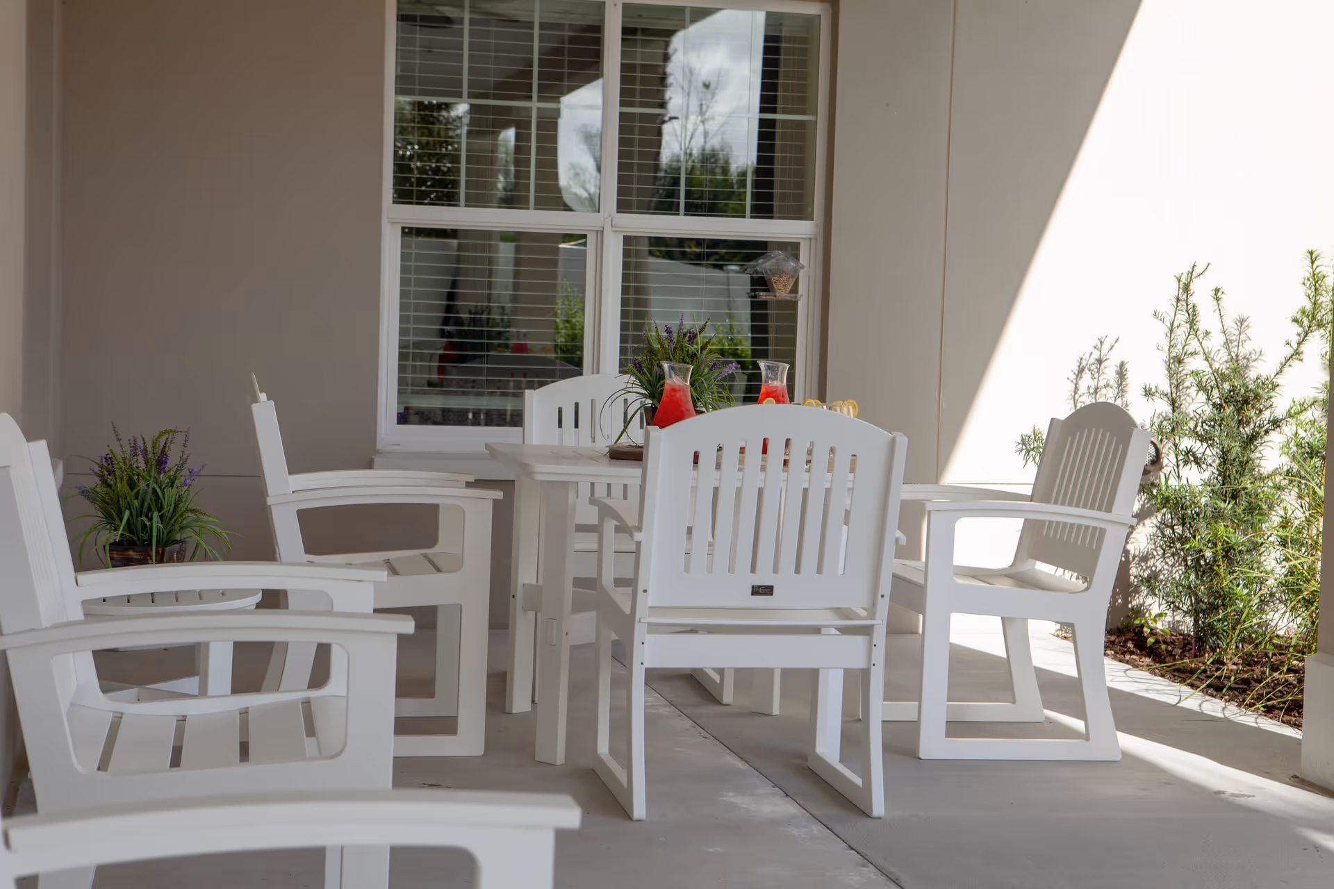 Outdoor patio area with white wooden chairs and a table. The table has two pitchers of red beverage and a small plant. There are plants along the wall and a window with white blinds in the background.