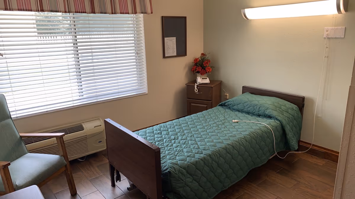 A small nursing home bedroom with a single bed covered in a green quilted blanket. Next to the bed is a wooden nightstand with a telephone and a vase of red flowers. A green armchair is positioned near a window with closed blinds. The room has wooden flooring and a wall-mounted light above the bed.