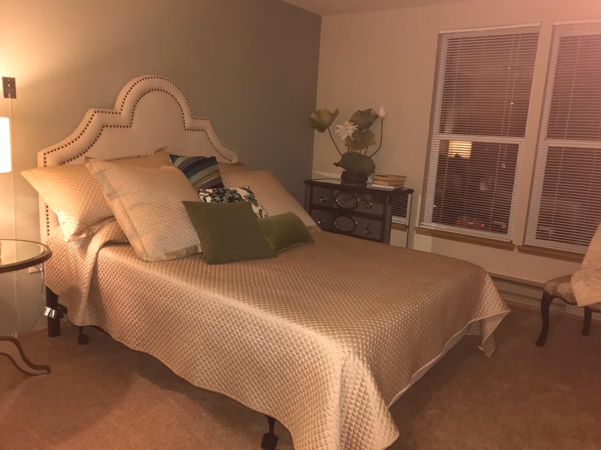 A neatly made bed with a beige quilted bedspread and multiple pillows, including green accent pillows, in a bedroom. The bed has a decorative upholstered headboard with nailhead trim. Next to the bed is a small round glass-top side table with a lamp, and on the other side is a dark wooden dresser with a vase of flowers and some books. Two windows with blinds are visible on the wall behind the dresser, and a chair with a light-colored throw is partially visible on the right side of the image.