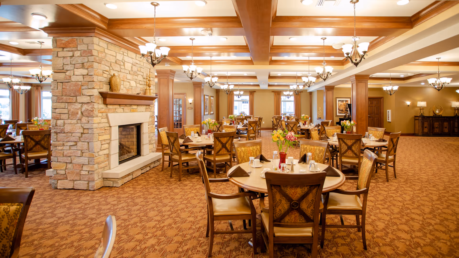 A spacious dining room in a senior living facility with multiple round tables set with glasses, napkins, and floral centerpieces. The room features warm lighting, a stone fireplace, wooden beams on the ceiling, and carpeted floors. Chairs with patterned upholstery surround the tables, and large windows allow natural light to enter.