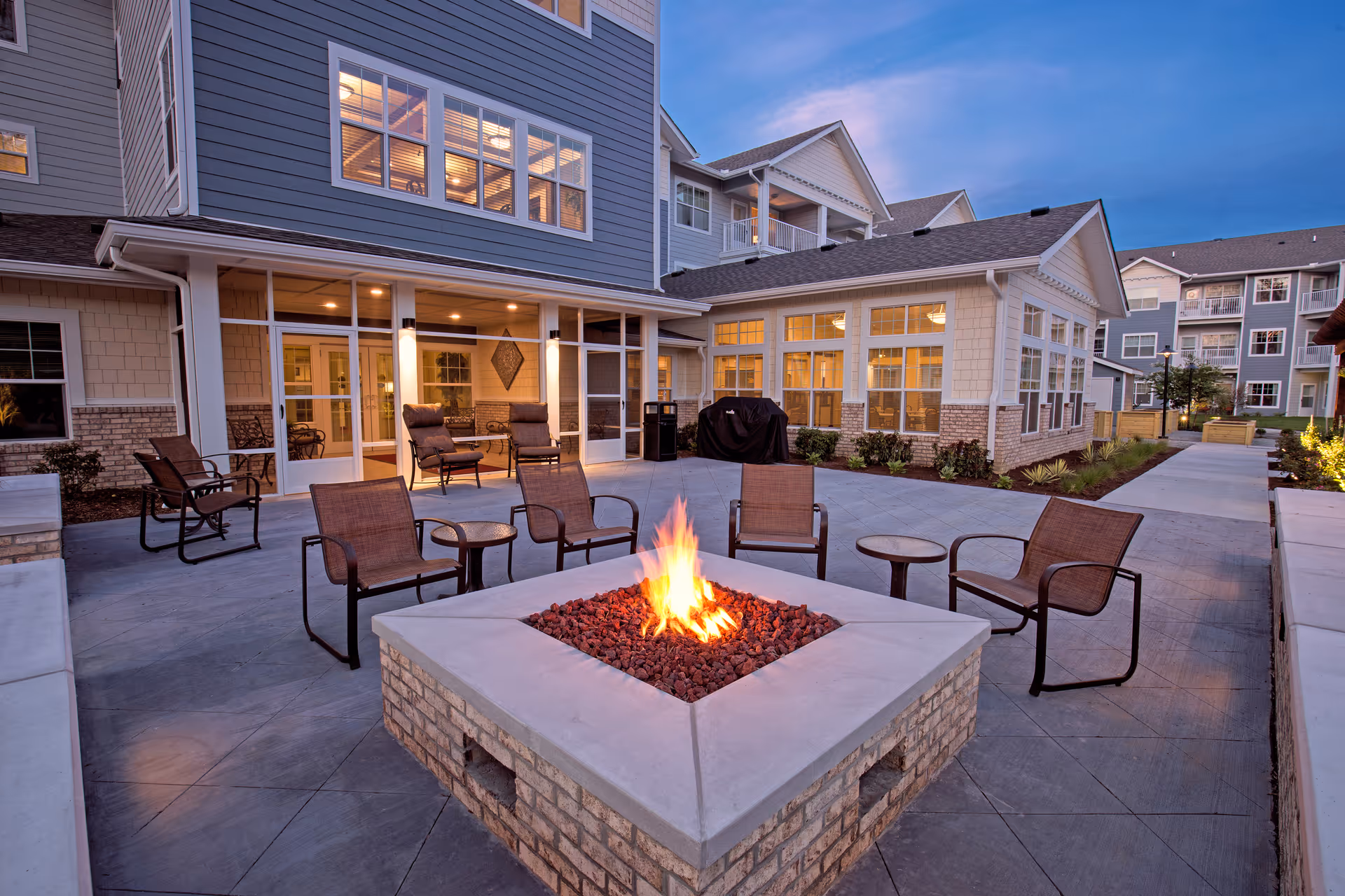 Outdoor courtyard patio with a central stone fire pit surrounded by chairs in front of a multi-story senior living building.