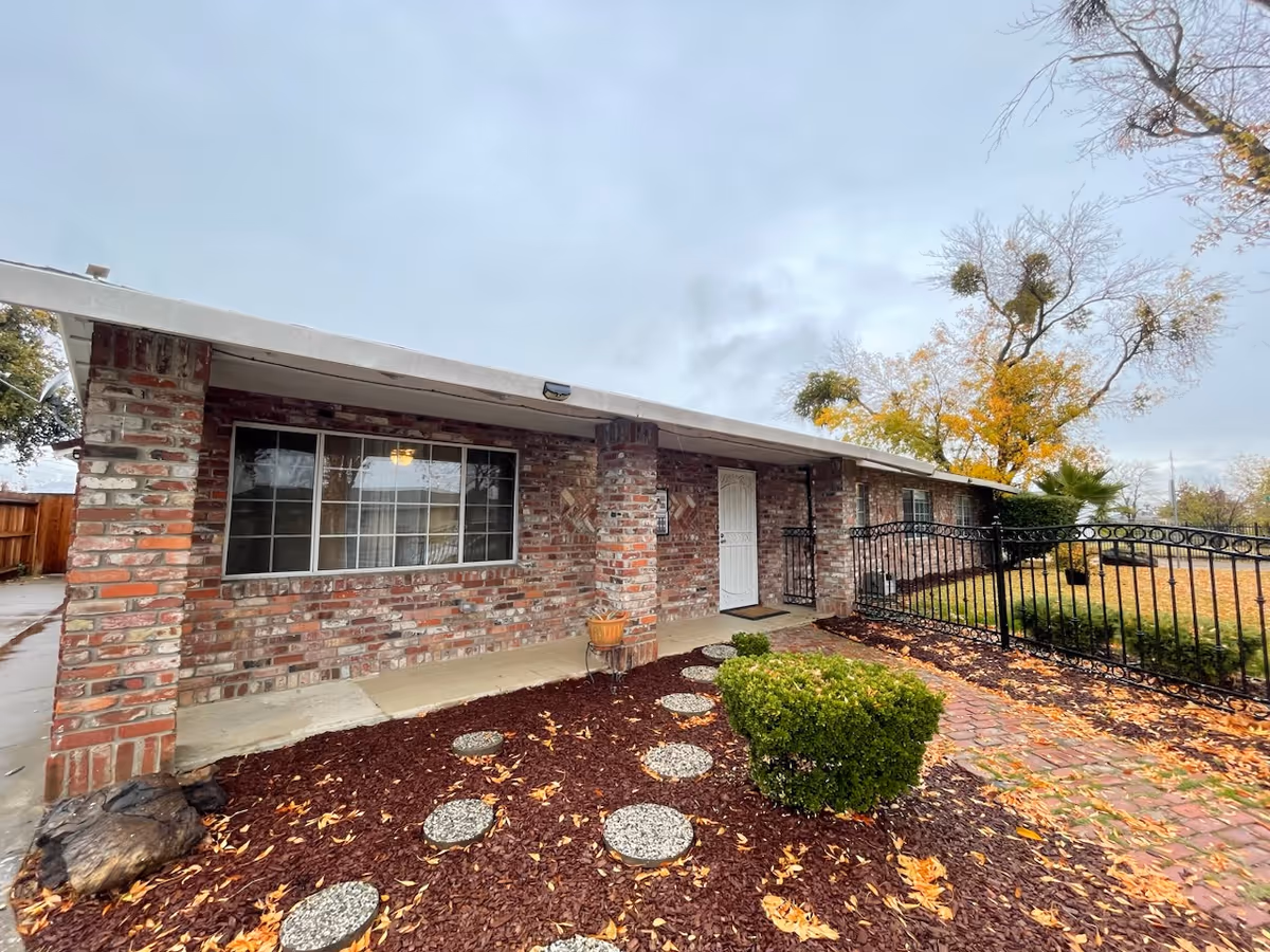 Exterior view of a single-story brick building with a covered porch, a white door, large window, and a small landscaped garden with stepping stones and bushes. There is a black metal fence on the right side and trees with autumn leaves in the background under a cloudy sky.