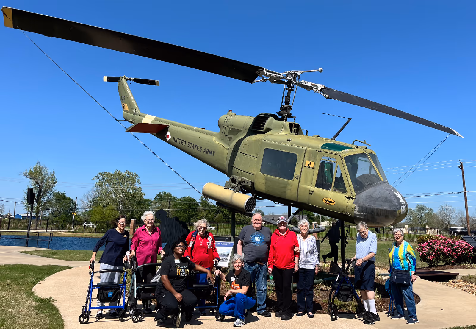 A group of elderly people and two caregivers posing in front of a green United States Army helicopter displayed outdoors on a sunny day with a clear blue sky. The group is standing and sitting on a paved path surrounded by grass and some bushes.
