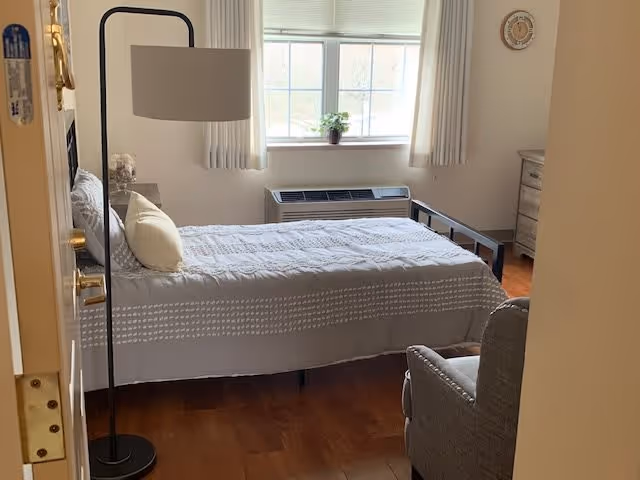A neatly made single bed with a gray and white patterned bedspread and a light yellow pillow in a bright bedroom. There is a tall black floor lamp next to the bed, a gray armchair in the foreground, a wooden dresser against the wall, a small plant on the windowsill, and a wall clock above the dresser. The room has wooden flooring and light-colored walls with a window letting in natural light.