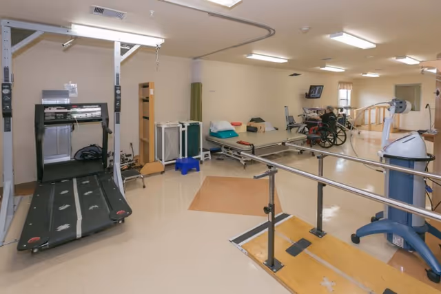 A rehabilitation therapy room with a treadmill, parallel bars, treatment tables, exercise equipment, and a person in a wheelchair in the background.