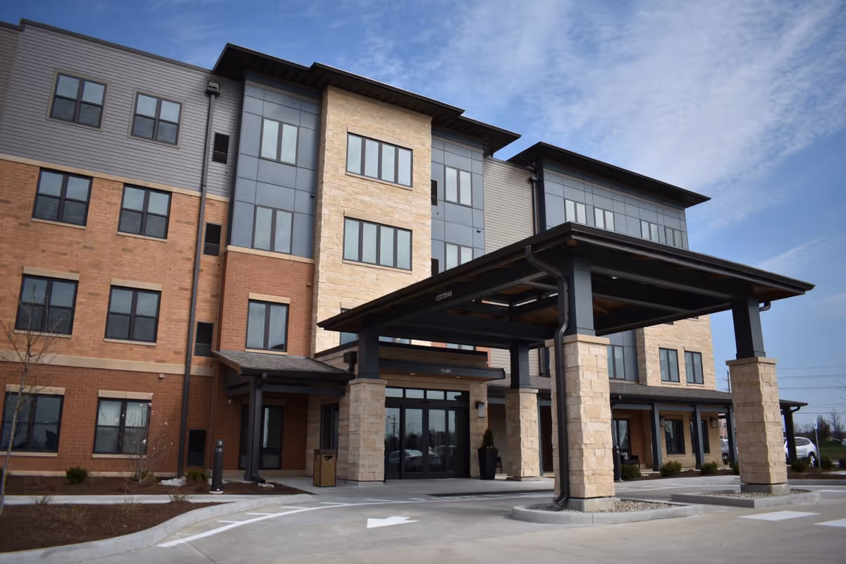 Exterior view of a modern multi-story senior living facility building with a covered entrance supported by stone pillars, large windows, and a clear sky in the background.