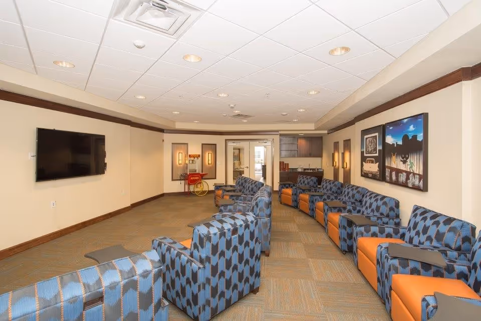A long common room with patterned blue and orange armchairs arranged in two rows facing a wall-mounted TV and artwork.