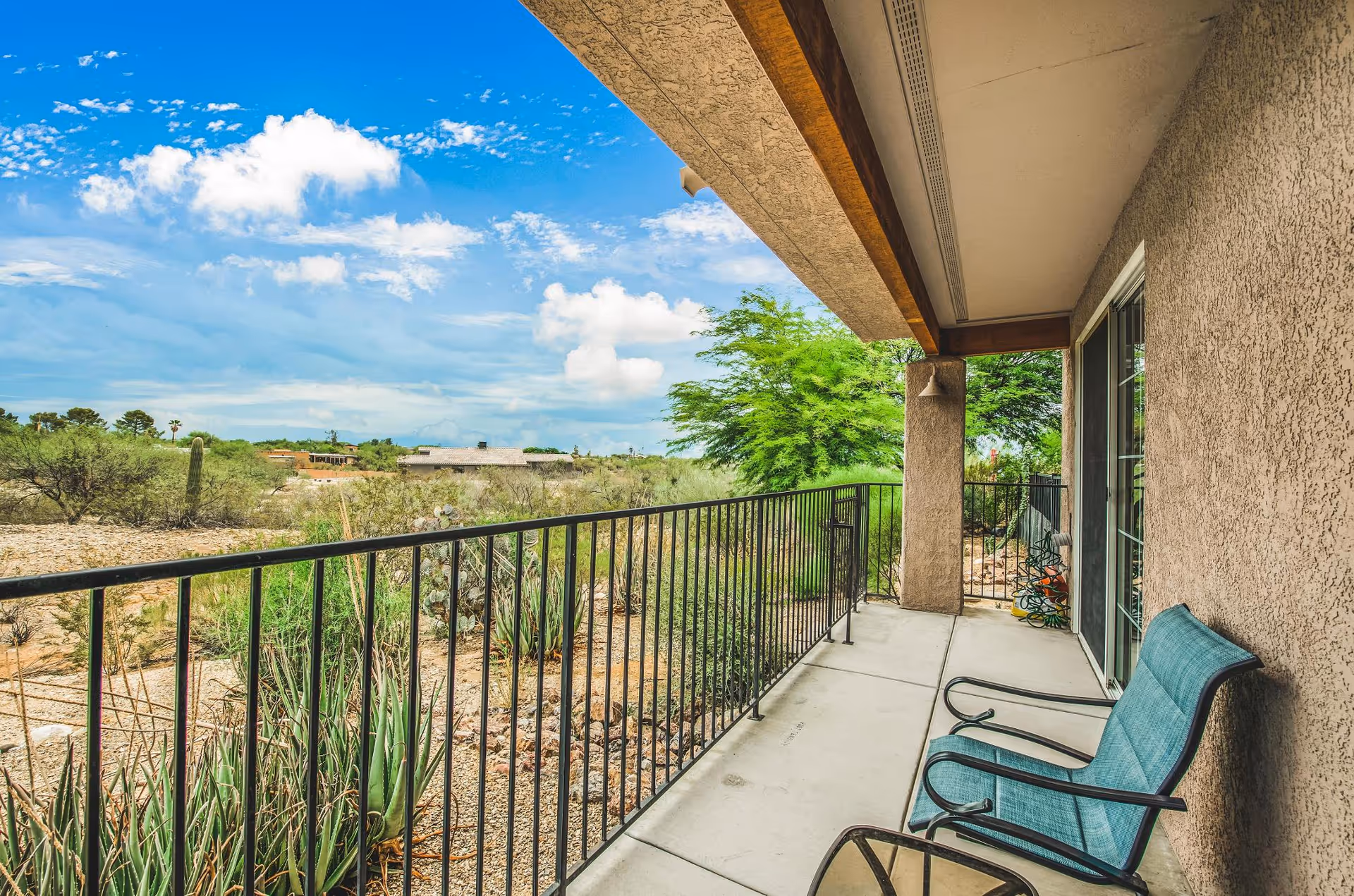A covered outdoor patio area with two blue cushioned chairs and a small glass-top table. The patio has a black metal railing and overlooks a desert landscape with cacti, shrubs, and distant buildings under a partly cloudy blue sky.