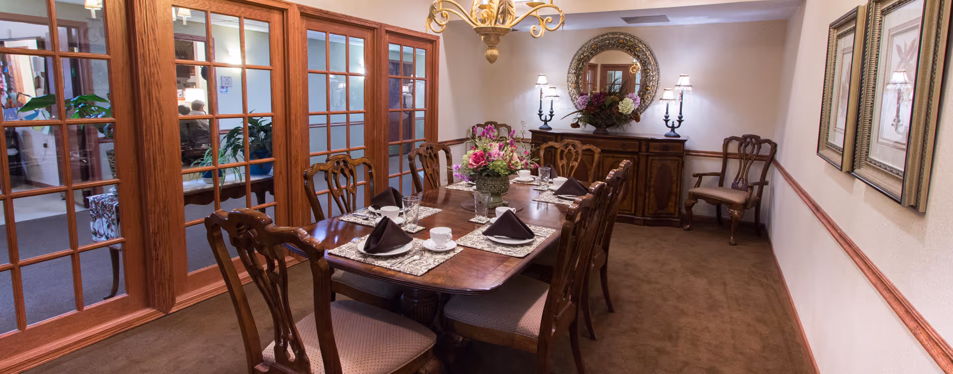 A formal dining room with a long wooden table set for six people, featuring folded napkins, glasses, and a floral centerpiece. The room has wooden framed glass doors on one side, a sideboard with a round mirror and decorative lamps on the far wall, and framed artwork on the right wall.