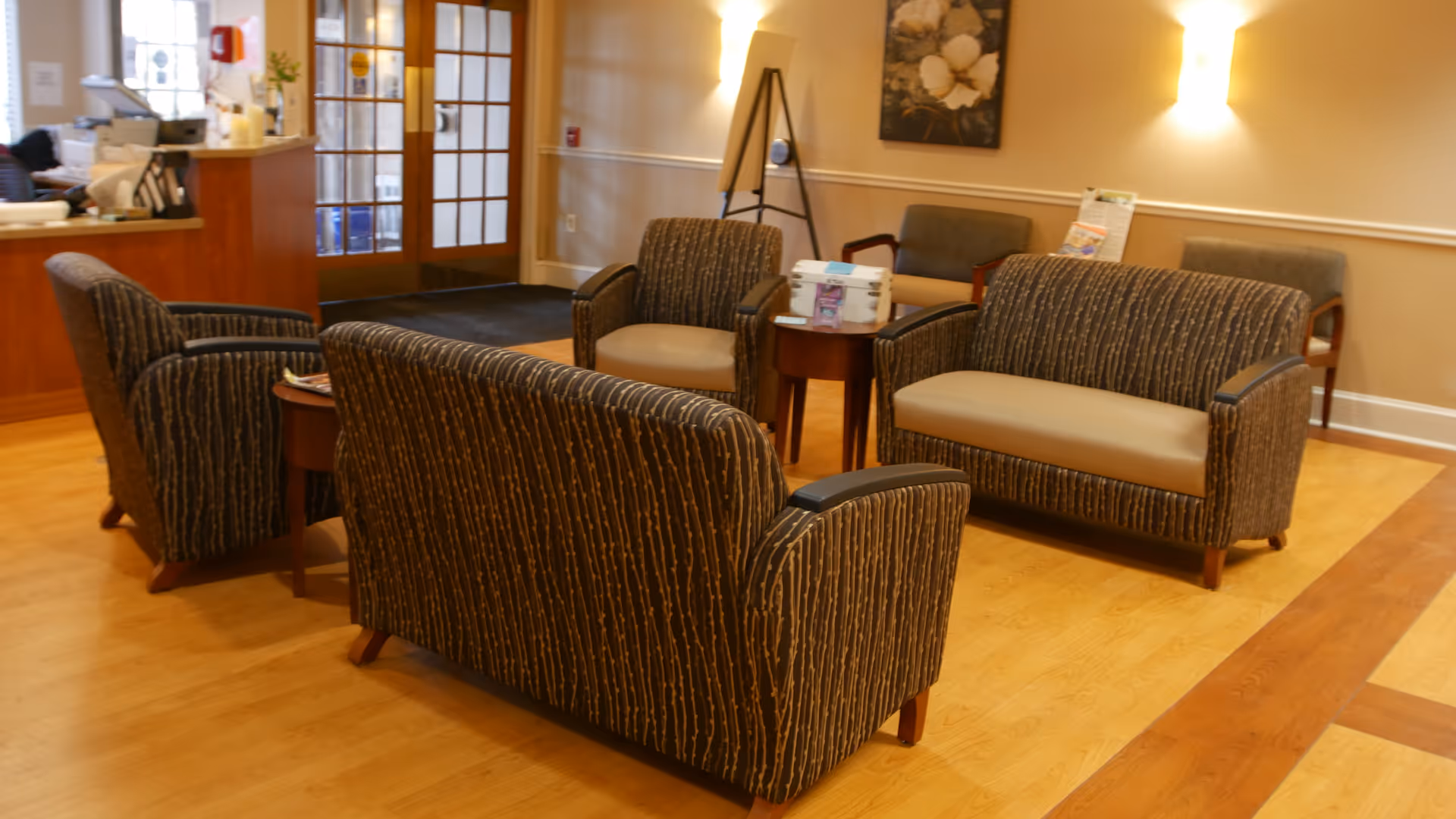 A cozy seating area in a nursing facility with patterned armchairs and sofas arranged around small wooden tables on a wood floor. The background shows a reception desk, a door with glass panels, wall art, and soft lighting.