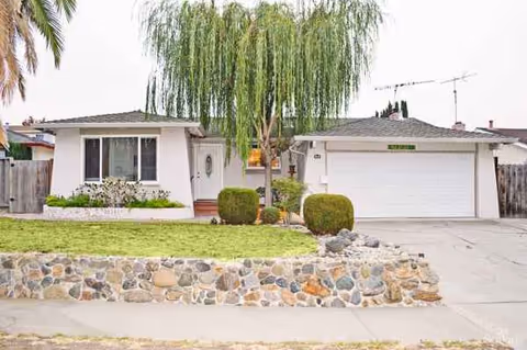 Single-story building with a two-car garage, front yard, stone wall, and palm trees.