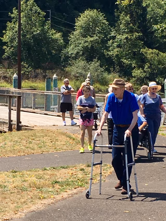 A group of elderly people walking outdoors on a paved path near a wooden bridge and green trees. One elderly man in the foreground is using a walker and wearing a blue shirt and a brown hat. Other people, including some using wheelchairs, are walking behind him.
