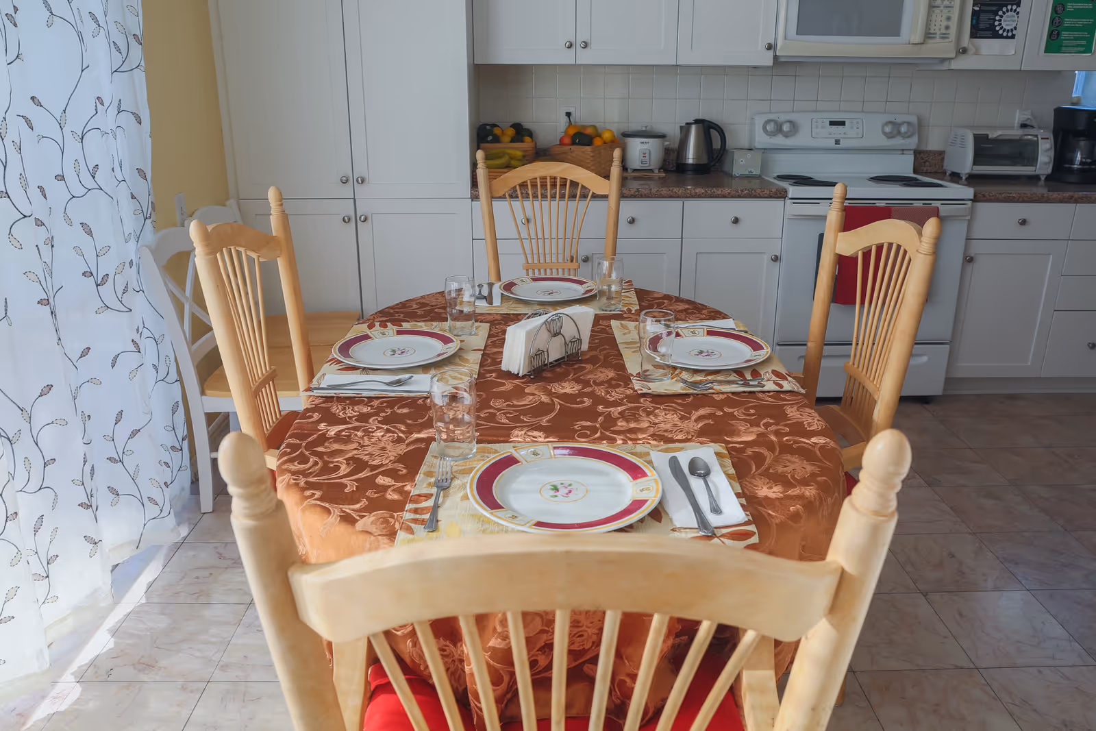 A dining table set for four with plates, glasses, and cutlery in a kitchen area. The table has a brown patterned tablecloth and wooden chairs with red cushions. In the background, there are white kitchen cabinets, a stove, a toaster, a kettle, and a basket of fruit. Sheer curtains with a leaf pattern cover a window to the left.