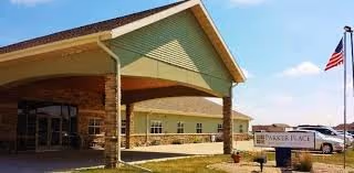 Exterior view of Parker Place Retirement Community building with a covered entrance, stone pillars, and an American flag on a flagpole. The sky is clear and blue.