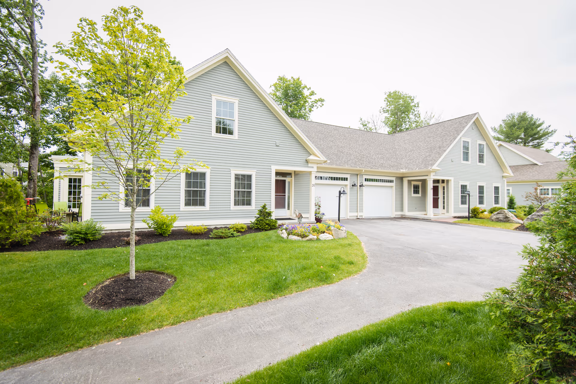 Exterior view of a light gray residential building with white trim, featuring multiple windows, two garage doors, and a driveway. The building is surrounded by green grass, small trees, shrubs, and a flower bed near the entrance. The sky is overcast.