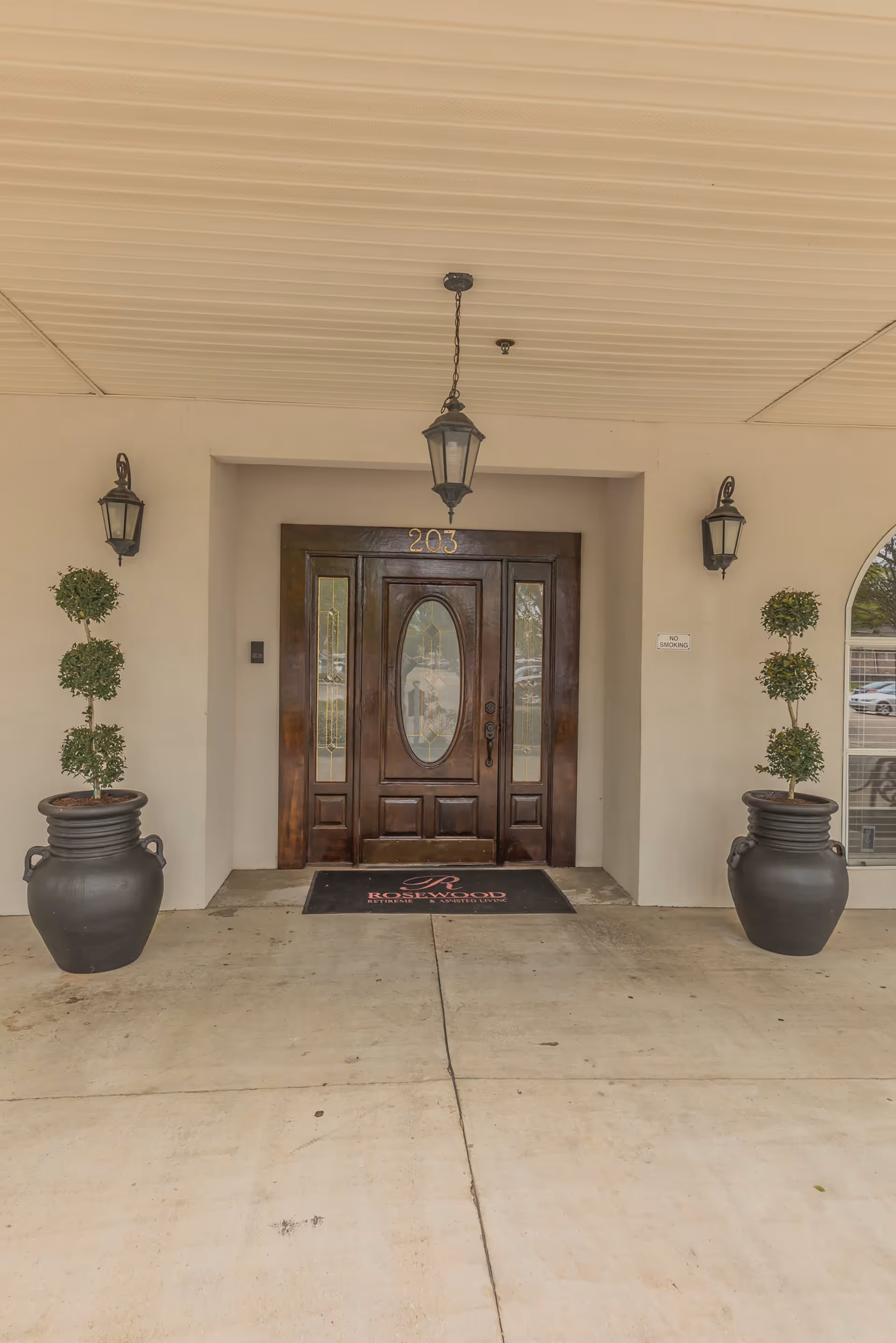 Entrance to Rosewood Assisted Living facility featuring a wooden door with glass panels, two large black planters with topiary plants on either side, a hanging lantern light above the door, and wall-mounted lantern lights on both sides.