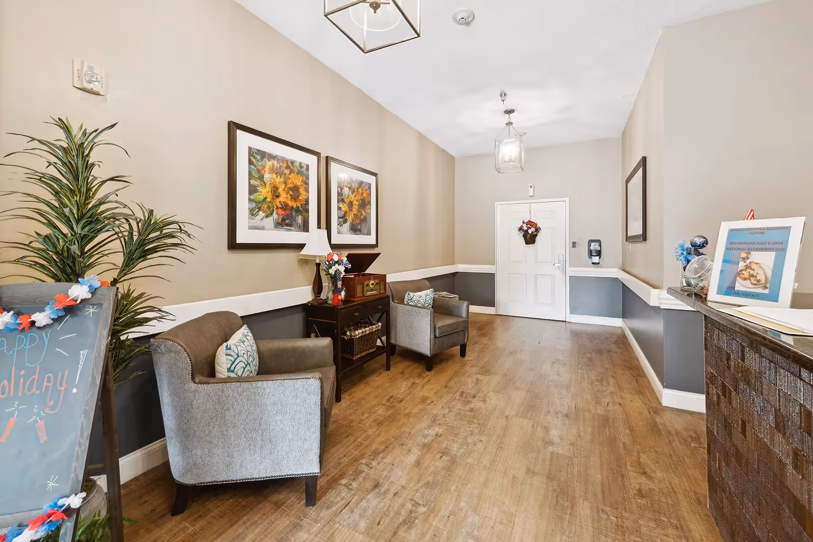 A hallway in a memory care community with wooden flooring and beige walls. Two gray armchairs with patterned pillows are placed along the left wall near a small table with a lamp and decorative items. Above the chairs are two framed pictures of sunflowers. A potted plant and a chalkboard sign with holiday decorations are also visible. At the end of the hallway is a white door with a floral wreath and a hand sanitizer dispenser mounted on the wall. On the right side, there is a counter with a framed sign and some decorative items.