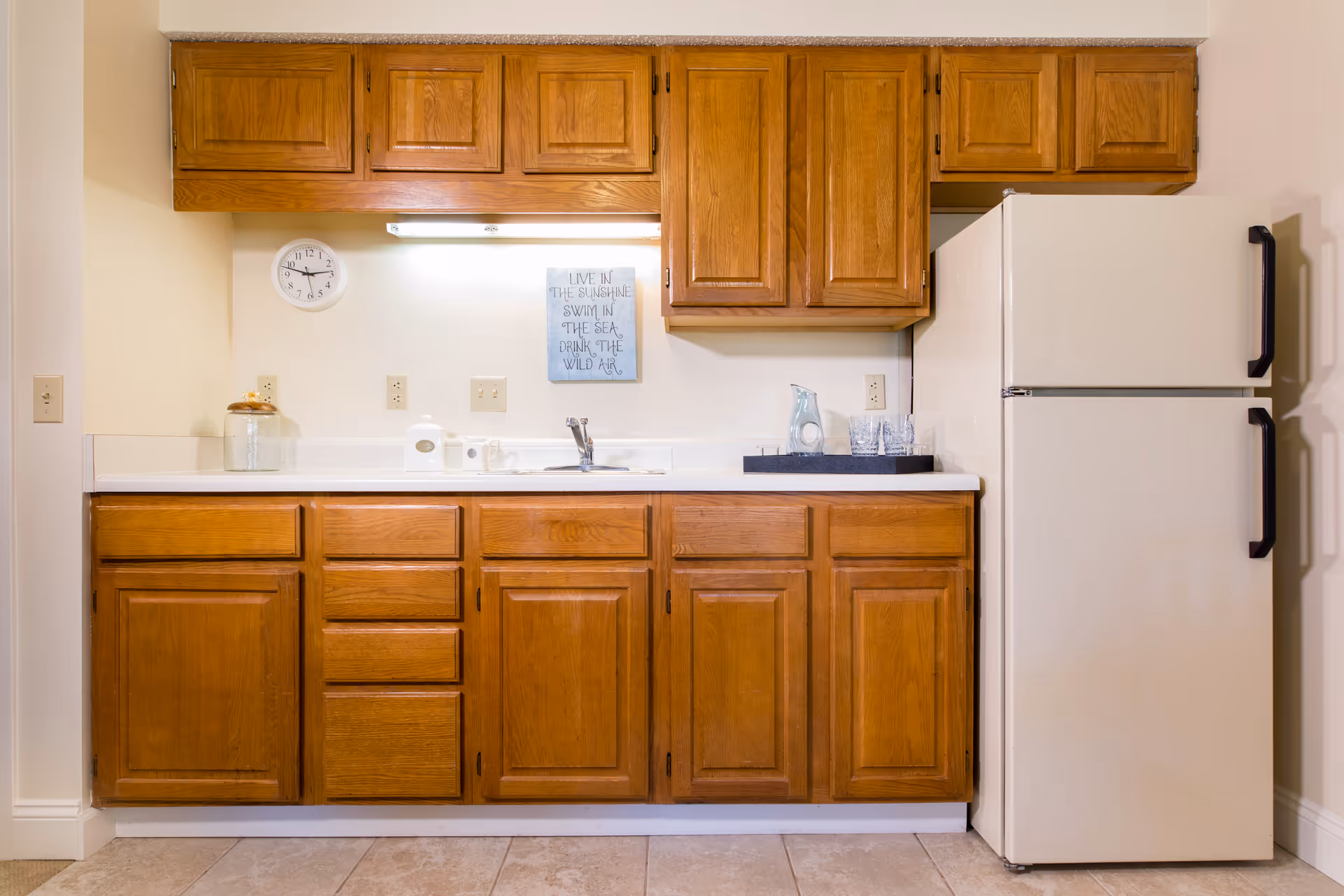 A kitchen area with wooden cabinets above and below a white countertop. There is a white refrigerator on the right side, a sink in the center of the counter, a clock on the wall to the left, and a small decorative sign above the sink. A tray with a pitcher and glasses is placed on the counter near the refrigerator.